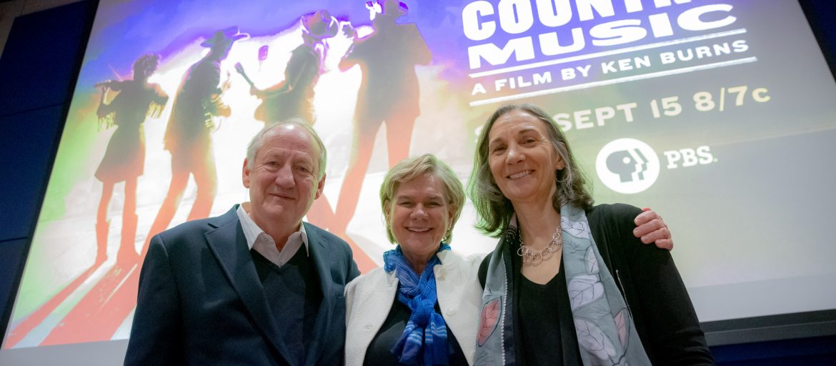 Three people smiling in front of the Country Music documentary image.