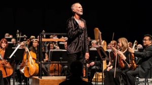 Composer John Luther Adams stands before an orchestra facing the audience with hand on heart