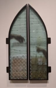 Work has appearance of church glass panels, and shows two hands with fingers reaching toward each other against a backdrop of barbed wire and overcast skies. The hands are separated by the division between the two panels.