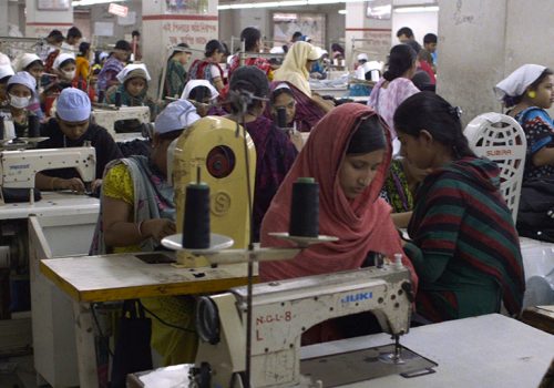 Crowded room full of people at sewing machines, many wearing face masks
