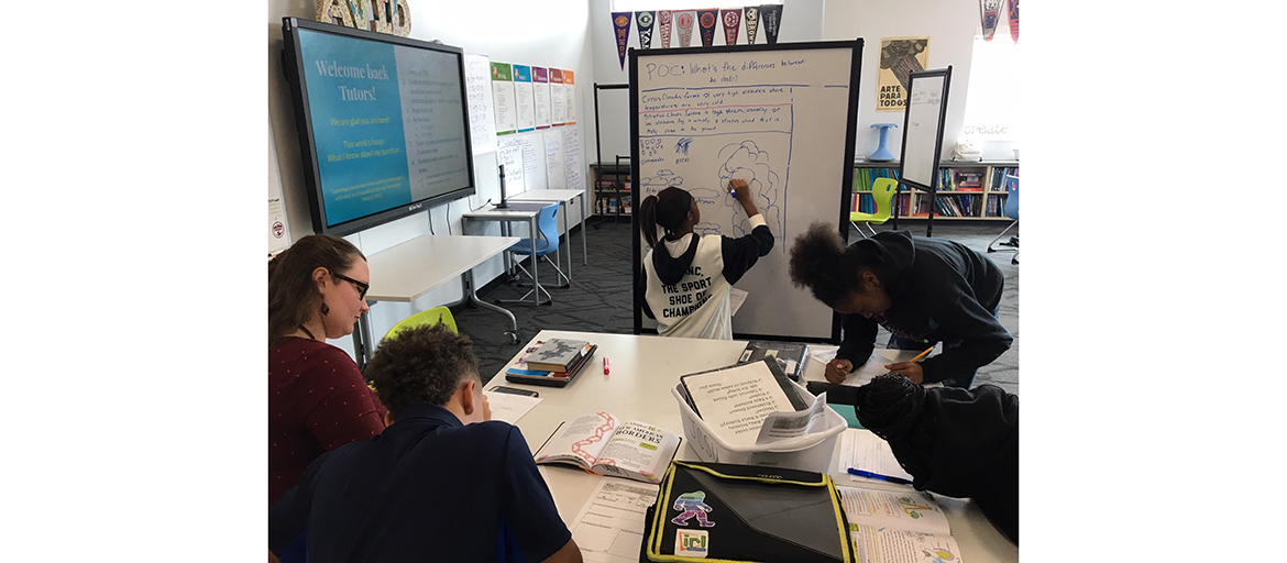 middle school students and tutor sit around a table while one student writes on a white board