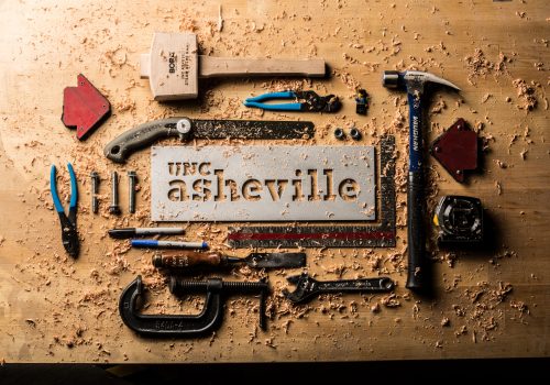 tools and sawdust surrounding a metal sign with CNC cut letters reading UNC Asheville