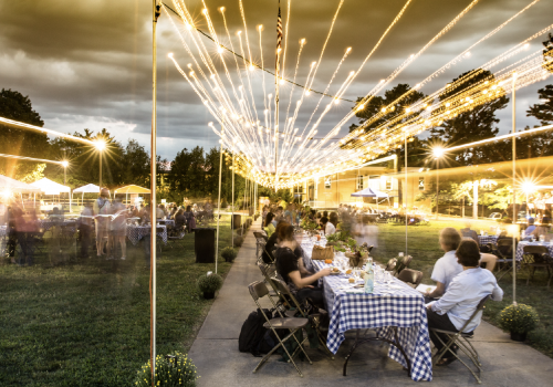 Wide shot of the farm to table dinner. Many people sitting around a long table, with string lights overhead