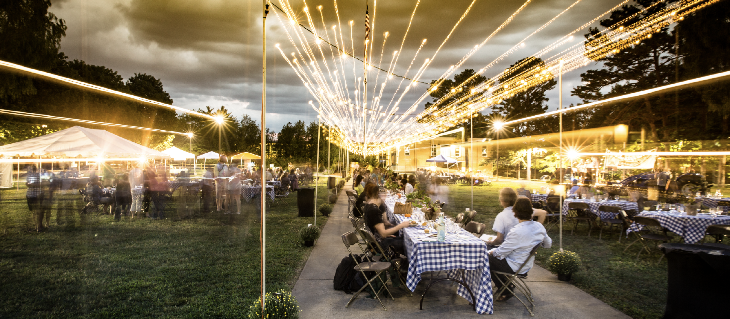 092822_farmtotable_v5_event-post_1170x512 Wide shot of the farm to table dinner. Many people sitting around a long table, with string lights overhead