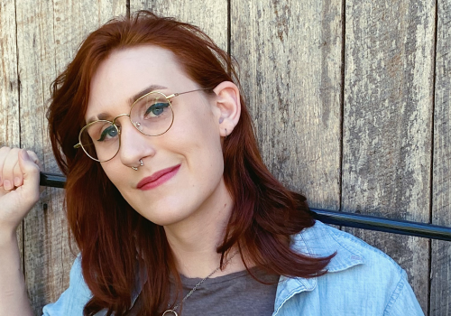 Headshot of Torrin A. Greathouse, holding a cane behind their neck, casually leaning against a fence.