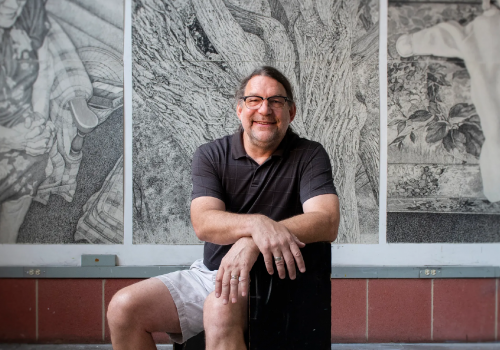 Artist Gerry Wubben smiling, posing in front of three of his works of art: large graphite drawings on display.