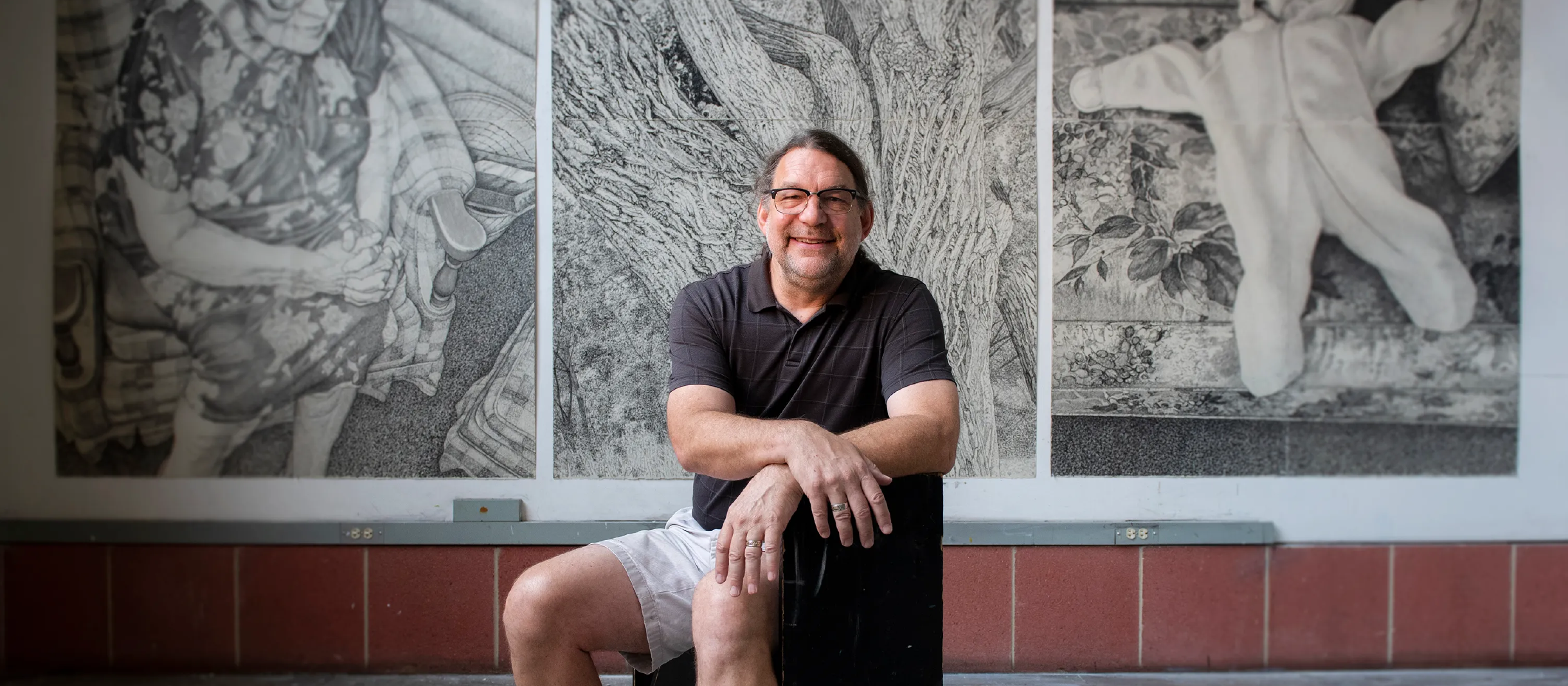 090522_GerryWuben_Art_v3_event-post_1170x512 Artist Gerry Wubben smiling, posing in front of three of his works of art: large graphite drawings on display.
