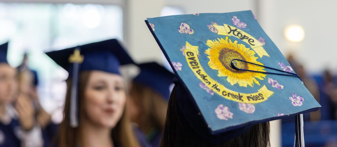 A student wearing a graduate's cap decorated to say "hope even when the creek rises"
