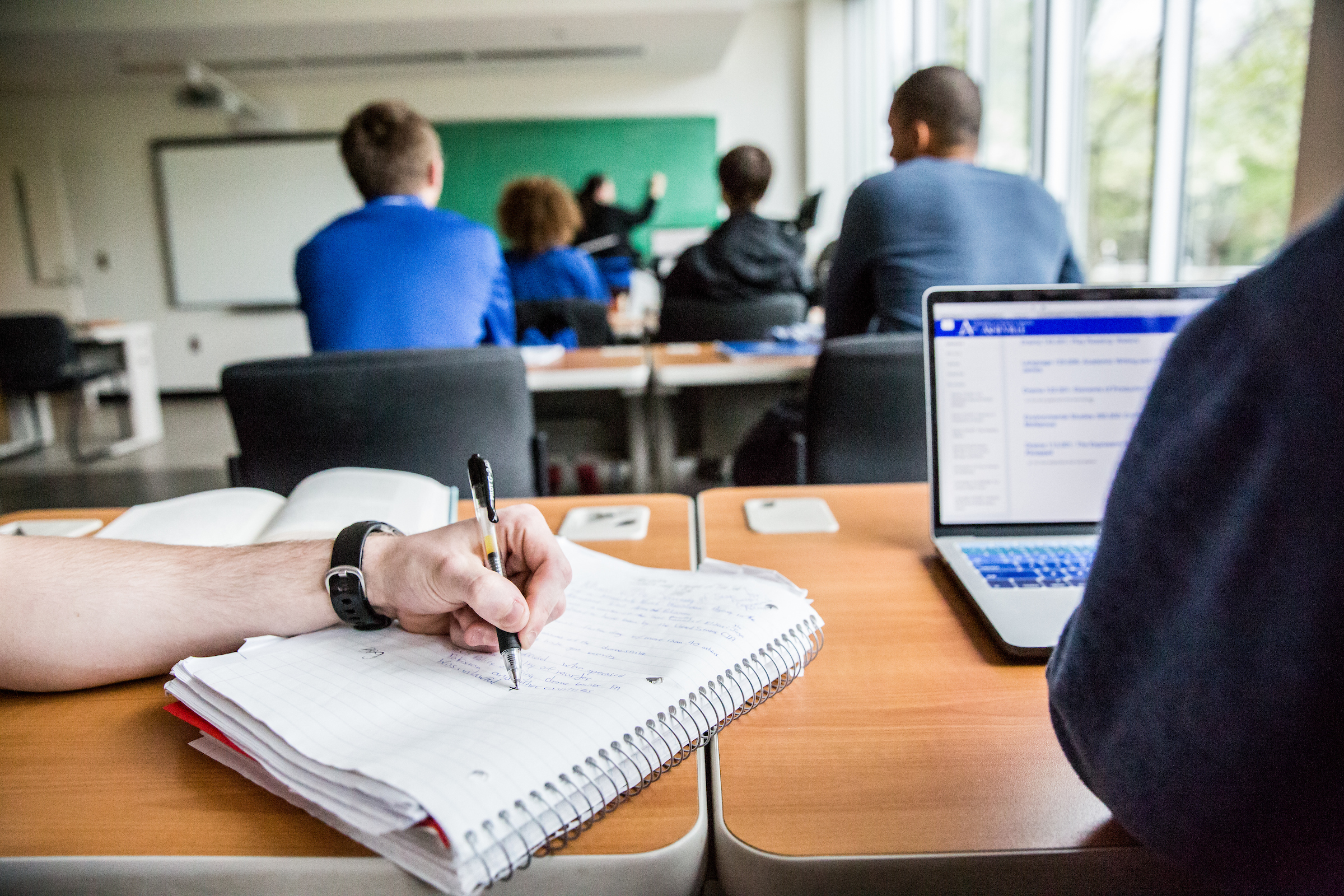 A close-up of a student taking notes while a professor lectures in the background.