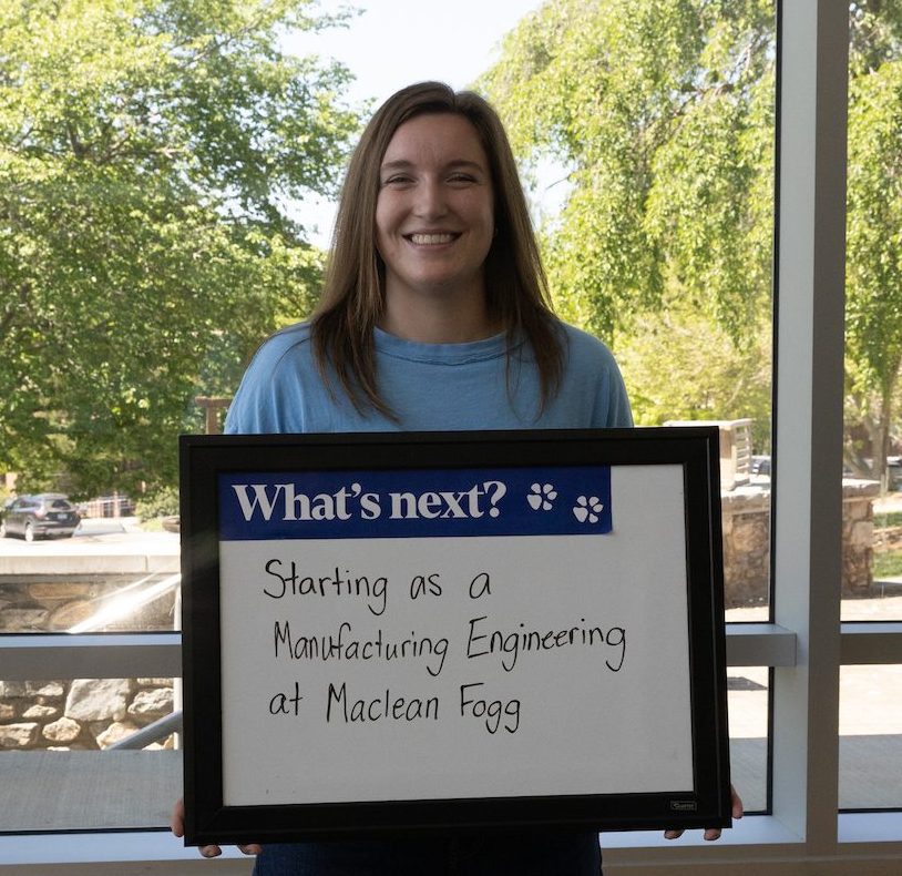 A person smiles while holding a sign that reads "What's next? Starting as a Manufacturing Engineering at Maclean Fogg," standing in front of a large window with trees visible outside.
