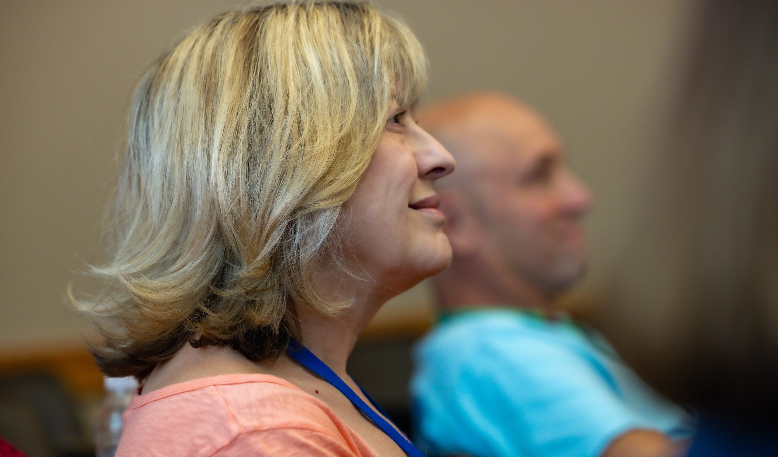 A smiling middle-aged woman with blond hair sits in a classroom with other older adults.