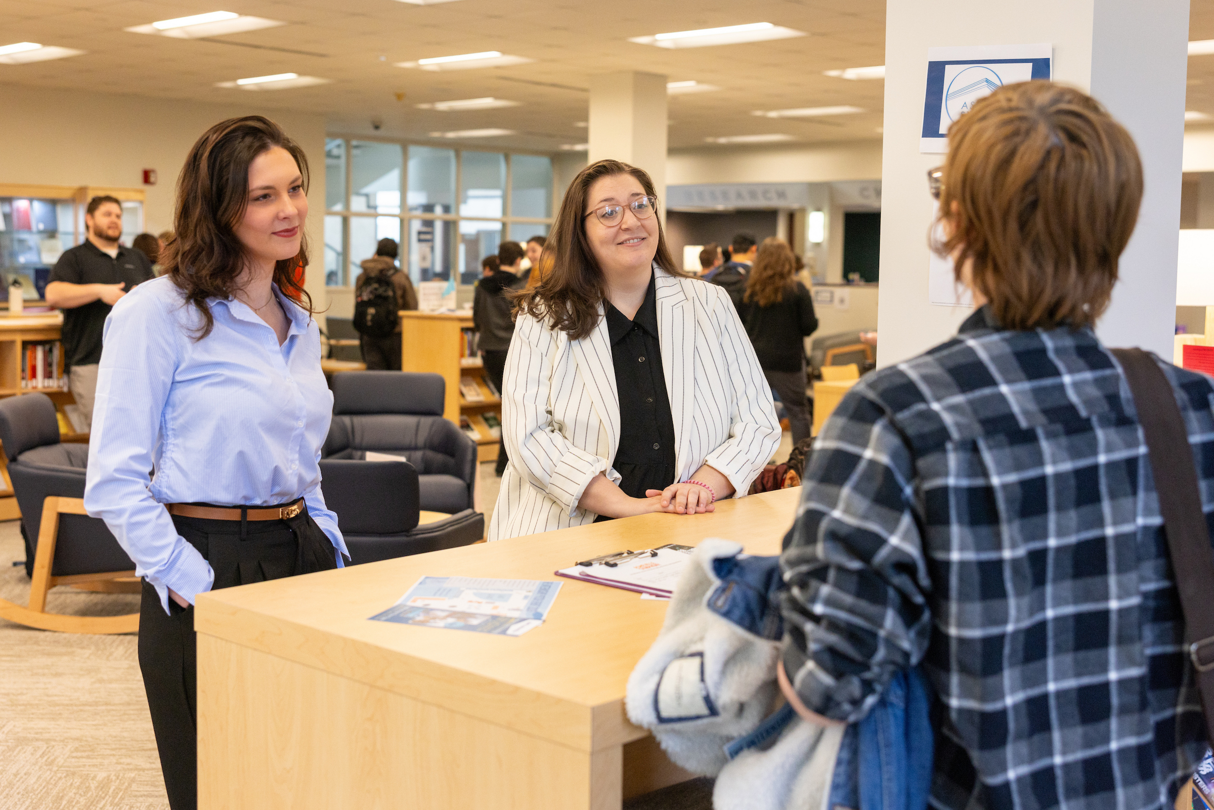Two women dressed business casual listen to a business pitch from someone wearing a plaid shirt.